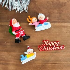 Retro set of vintage 1980s plastic Christmas cake toppers including Santa carrying sack, children on a sledge, and Happy Christmas motto.⁠